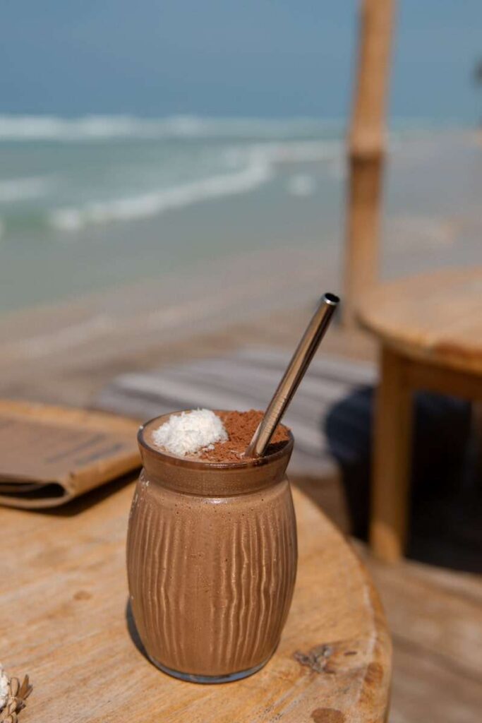A glass of chocolate smoothie topped with whipped cream and cocoa powder, served with a metal straw on a wooden table by the beachโa sweet treat to enjoy while exploring things to do in Ahangama, with the ocean and sky in the background.