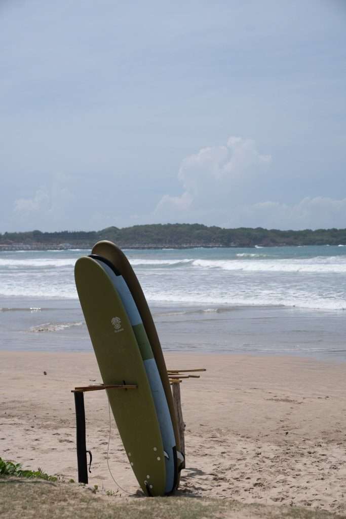 Two surfboards are propped up on a wooden stand on a sandy beach, with waves rolling in and a distant tree-covered coastline under a cloudy sky—perfect for those exploring Things To Do In Hiriketiya.