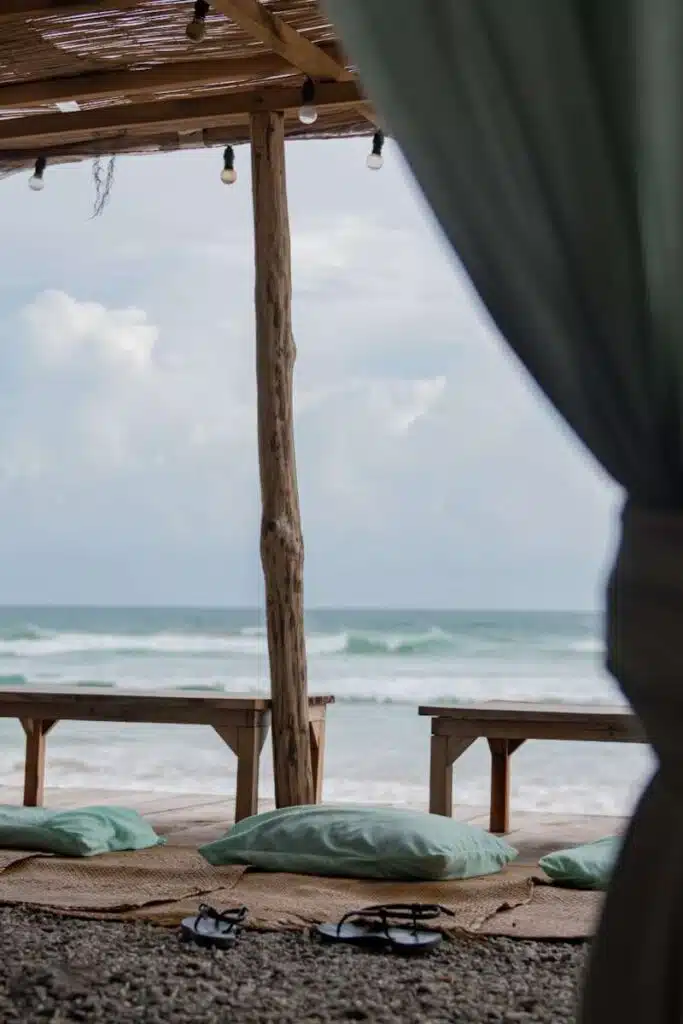 Outdoor beach seating area with mats and turquoise cushions under a wooden pergola, facing the ocean; sandals rest on the ground as a curtain frames the waves—perfect for relaxing and enjoying things to do in Ahangama.