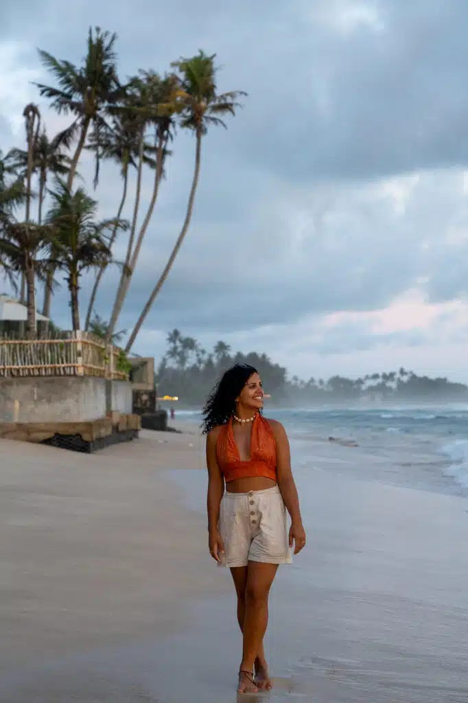 A woman in a red crop top and beige shorts walks barefoot on a sandy beach at sunset, palm trees and waves behind her. She smiles, looking to the side—one of the many relaxing things to do in Ahangama.
