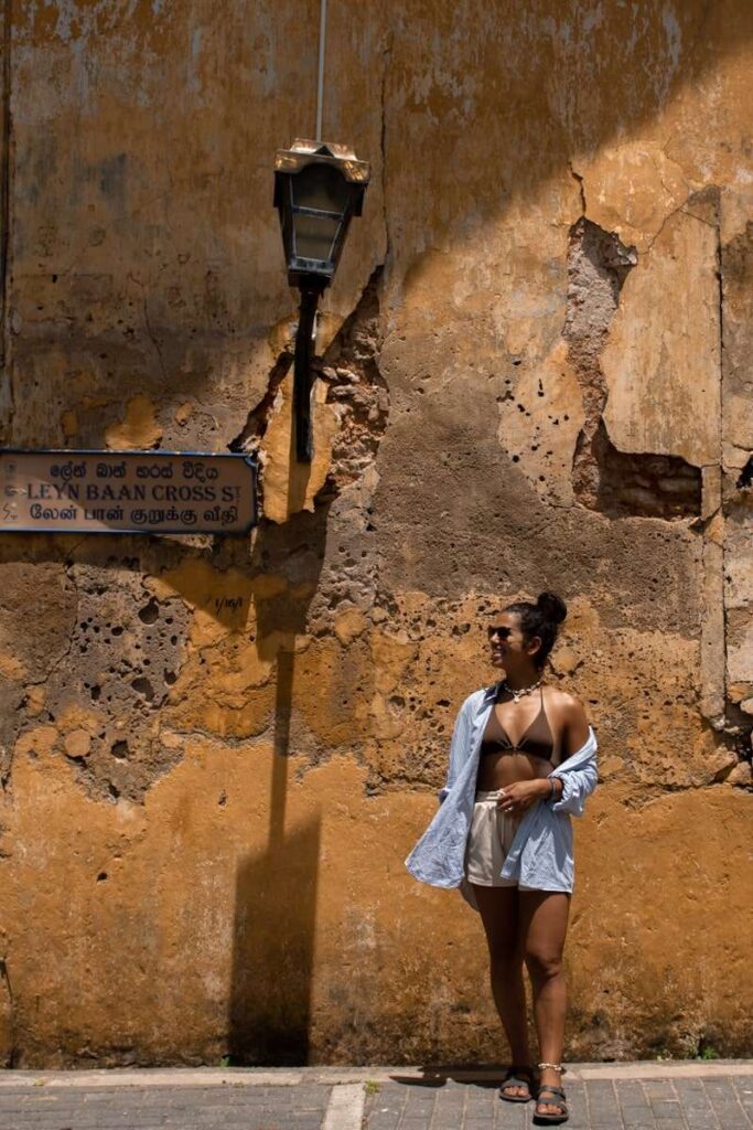 A woman in a bikini top, shorts, and open shirt stands under a streetlamp beside a weathered yellow wall with peeling paint and a street signโcapturing the laid-back vibe among the many things to do in Ahangama.