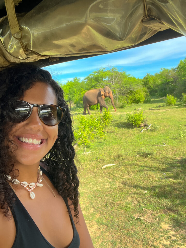 A woman wearing sunglasses and a shell necklace smiles for a selfie in a safari vehicleโone of the unforgettable things to do in Ahangamaโwith a large elephant on grassy land and trees under a blue sky in the background.