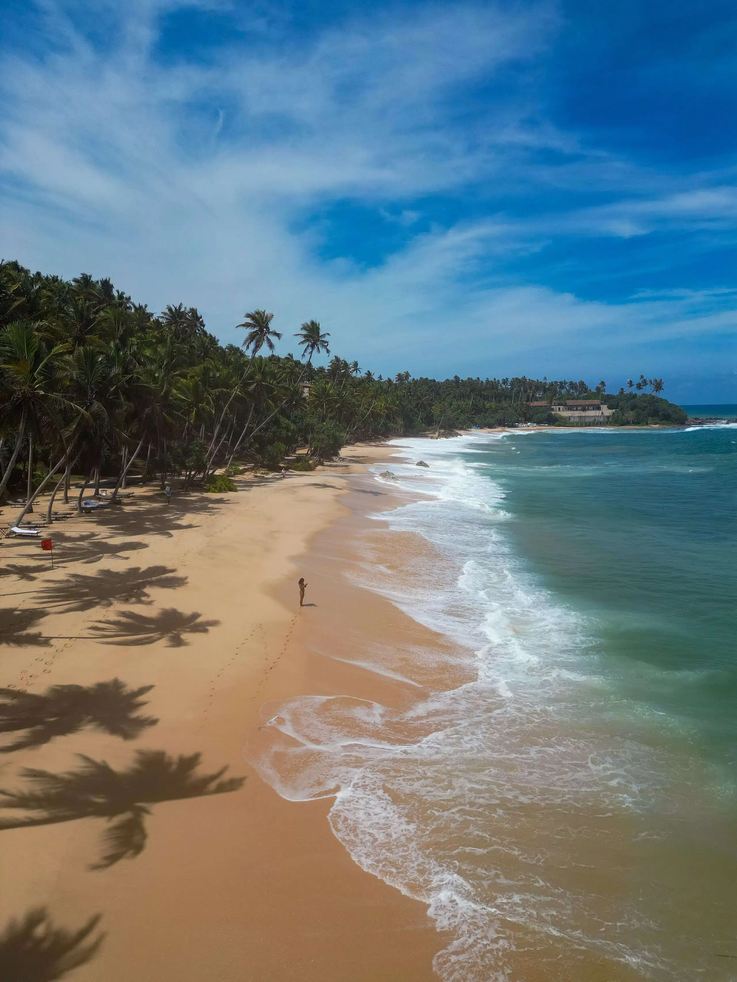 A person stands on a deserted tropical beach in Hiriketiya, bordered by tall palm trees on one side and the ocean on the other. The sky is clear with wispy clouds, and gentle waves wash onto the golden sand—a perfect start to exploring things to do in Hiriketiya.