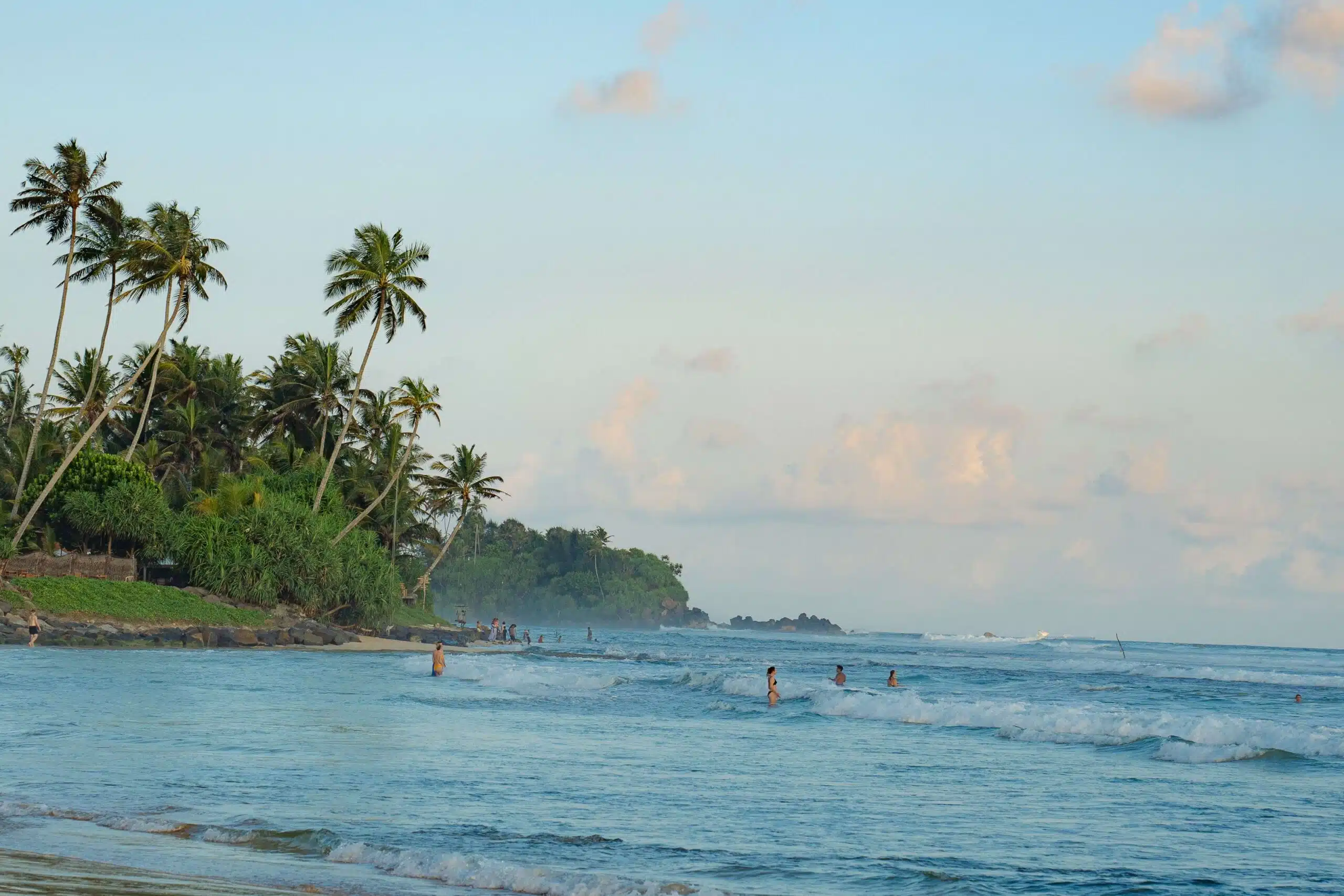 Tropical beach scene in Ahangama with tall palm trees lining the shore. People are swimming and playing in the gentle waves, under a blue sky with scattered clouds. A distant, tree-covered hill enhances the backdrop of this charming haven.