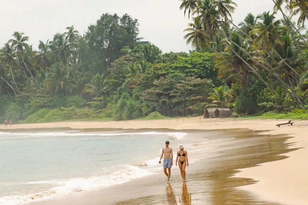 A man and a woman walk along a sandy tropical beach beside the ocean, exploring one of the best things to do in Hiriketiya, with tall palm trees and dense green foliage under a slightly overcast sky.