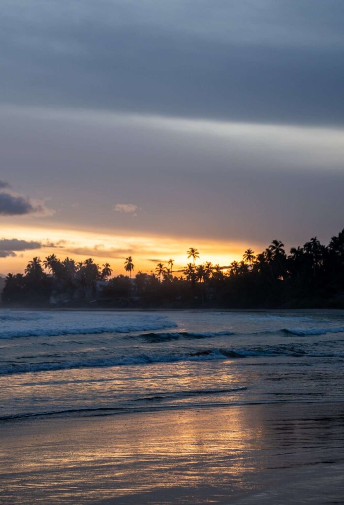 A serene beach at sunset with gentle waves, palm trees silhouetted against the colorful sky, and golden sunlight reflecting on wet sand—perfect for those seeking peaceful things to do in Hiriketiya.