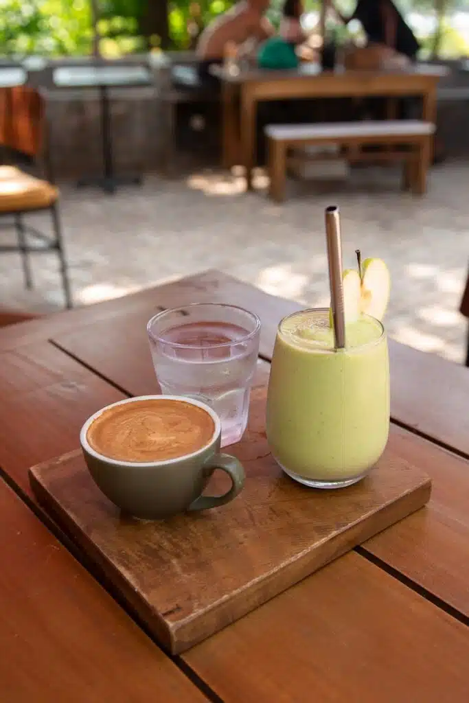 A cappuccino, a glass of water, and a green smoothie with apple slices and a metal straw sit on a wooden board atop a table in an outdoor cafe—perfect for relaxing after exploring Things To Do In Hiriketiya.