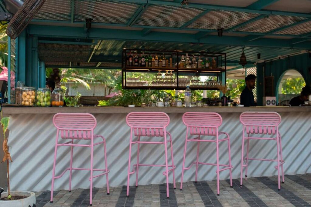 Outdoor bar with four pink metal stools lined up at a counter. Bottles and glasses rest on shelves behind the bar. Lush plants and sunlight hint at a tropical vibe—perfect for your list of Things To Do In Hiriketiya.