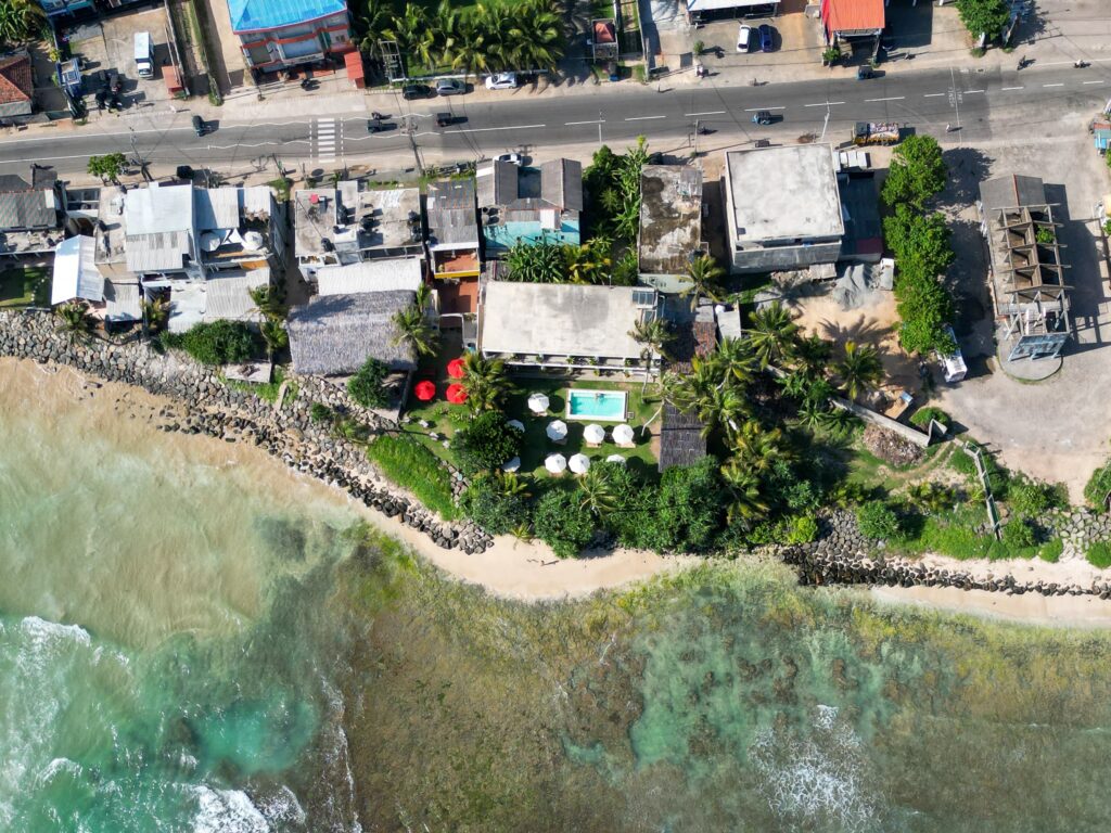 Aerial view of a coastal area with buildings and a road along the shoreline, a pool with white umbrellas, lush greenery, and waves washing onto the sandy beach edged by rocksโperfect for exploring the many things to do in Ahangama.