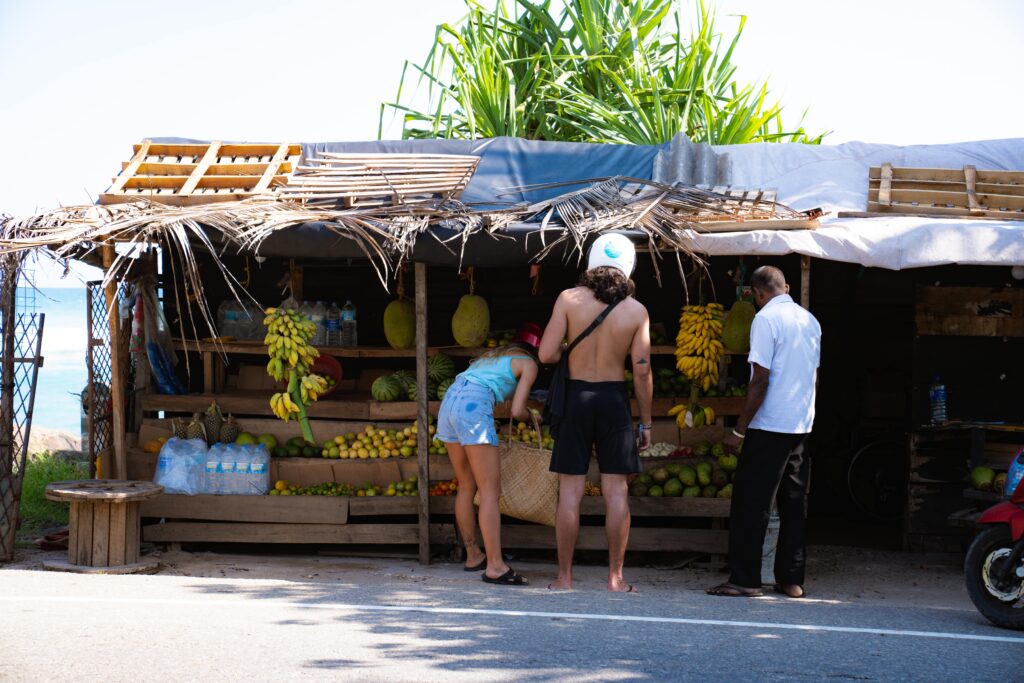 Two people browse fruits at a small outdoor wooden stand shaded by palm leaves, as a vendor stands nearby. Bananas, water bottles, and various fruits are displayedโa classic scene among the things to do in Ahangama.