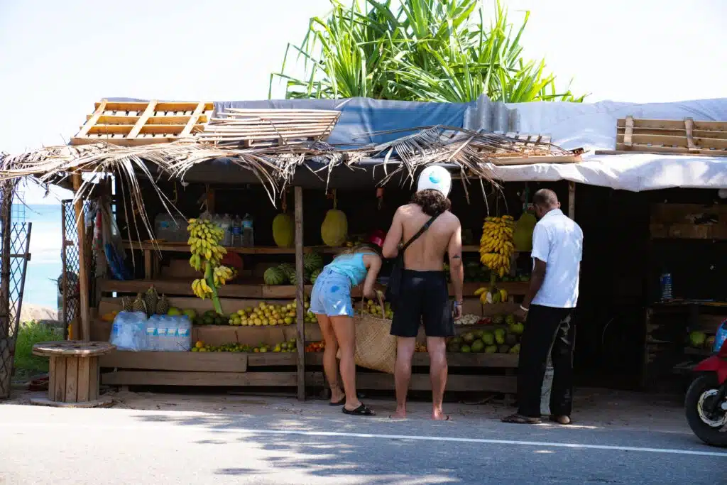 Two people browse fruits at a small outdoor wooden stand shaded by palm leaves, as a vendor stands nearby. Bananas, water bottles, and various fruits are displayed—a classic scene among the things to do in Ahangama.