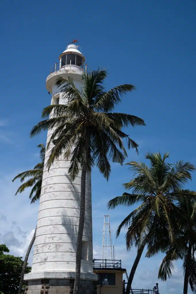 A tall white lighthouse stands beside swaying palm trees under a clear blue sky, making it one of the top things to do in Ahangama.
