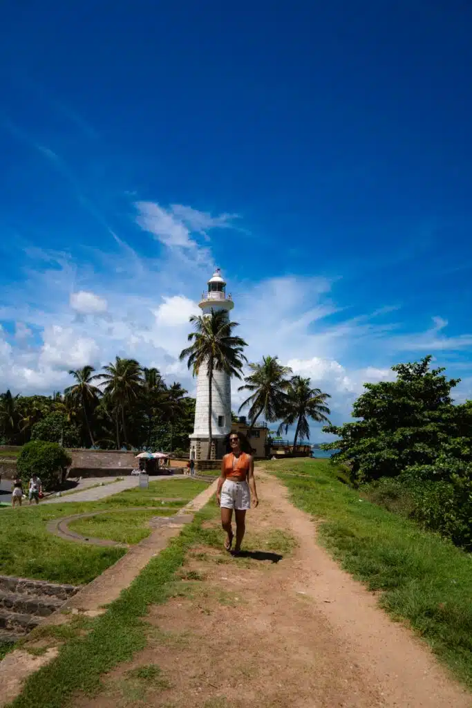 A person walks along a dirt path toward a white lighthouse surrounded by palm trees, under a bright blue sky with scattered clouds—one of the picturesque things to do in Ahangama.