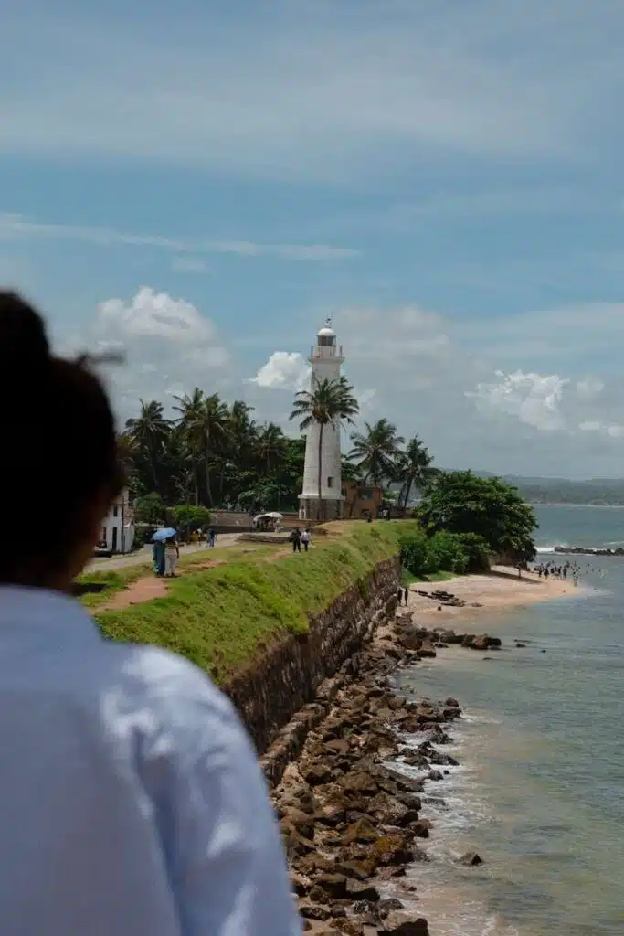 A person in the foreground looks toward a white lighthouse surrounded by palm trees on a rocky coastline—a perfect spot for those seeking things to do in Ahangama, with people walking along the path and ocean waves under a partly cloudy sky.