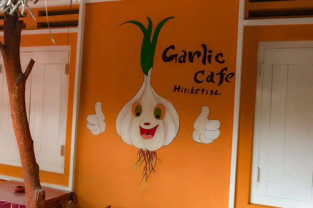 An orange wall features a cartoon garlic bulb giving thumbs up and smiling, with "Garlic Cafe Hiriketiya" text—one of the fun Things To Do In Hiriketiya. White doors frame this vibrant spot on either side.