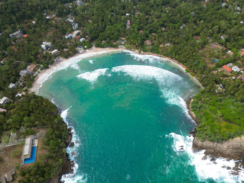 Aerial view of a crescent-shaped beach with turquoise water, white waves, green hills, and scattered houses nestled among dense trees—perfect for exploring the many things to do in Hiriketiya.