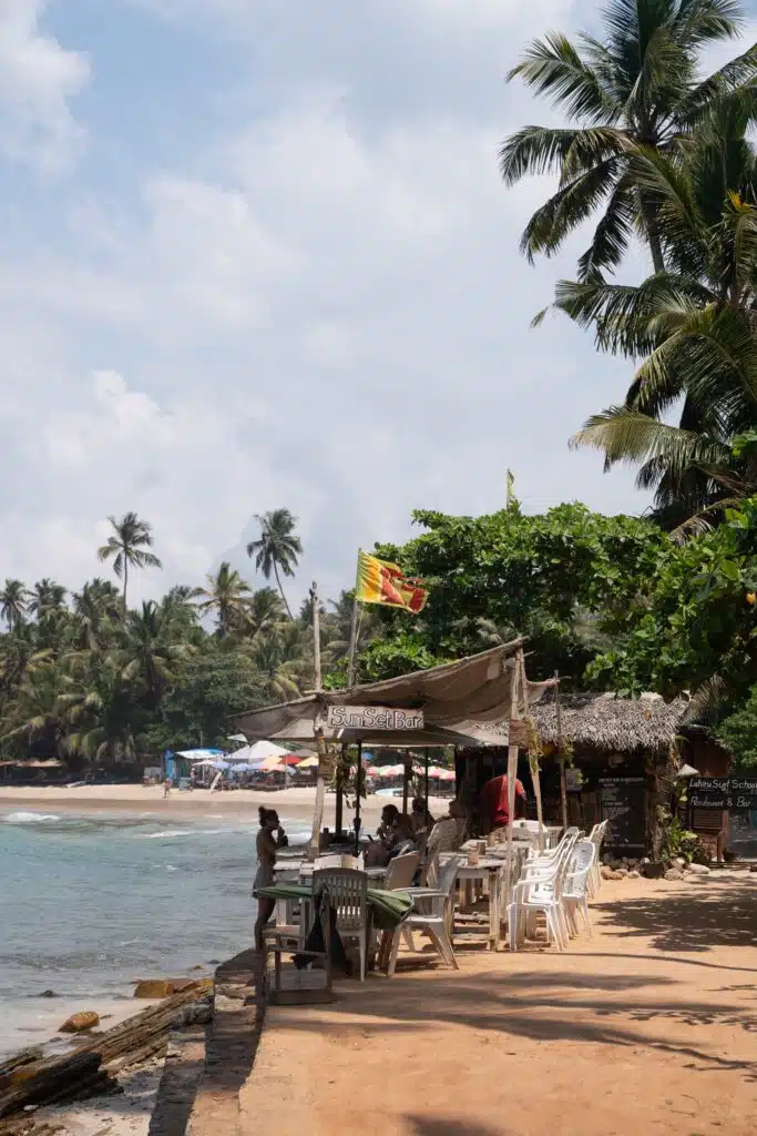 Outdoor beachside café with white plastic chairs and tables under a thatched canopy, surrounded by palm trees—a perfect spot to unwind while exploring Things To Do In Hiriketiya. The ocean and more palms provide a relaxing backdrop for visitors.