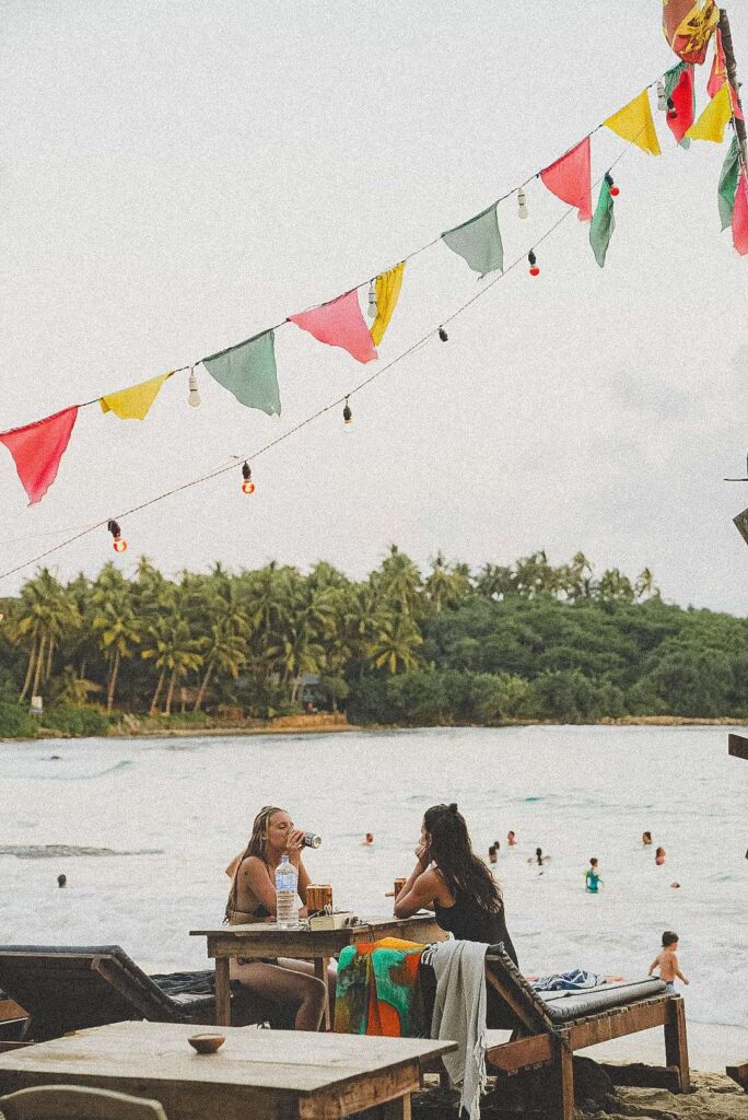 Two people sit at a wooden table on a beach, talking and enjoying drinks. Colorful flags and string lights hang above—a perfect scene if you’re searching for Things To Do In Hiriketiya, with swimming and palm-lined shores in the distance.