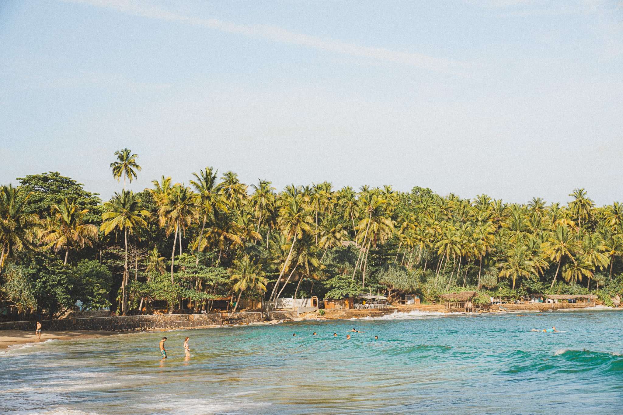 People swim and walk along the shore of a tropical beach lined with tall palm trees and dense greenery under a clear sky—one of the relaxing things to do in Hiriketiya.