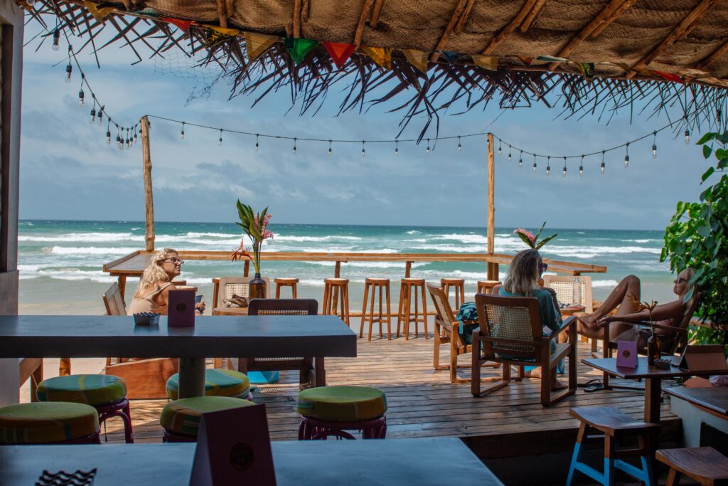 Outdoor beach bar with colorful stools and wooden tables, overlooking the oceanโa perfect spot to unwind. A few people relax on chairs, enjoying the scenic view. One of the top things to do in Ahangama under string lights and a thatched roof.