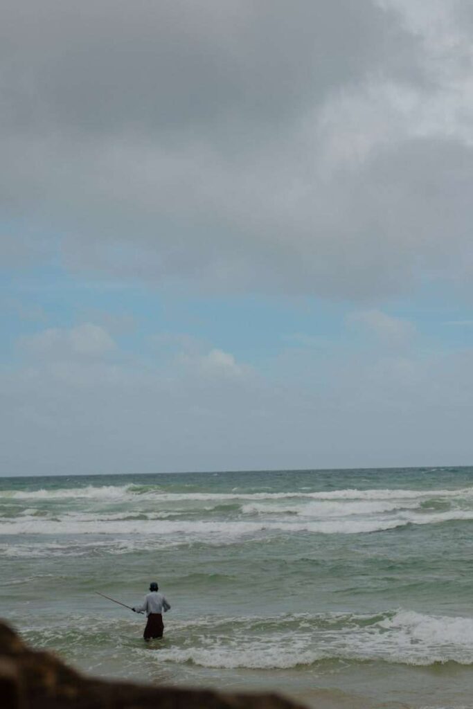 A person stands in shallow ocean water, holding a fishing rod under a cloudy sky with waves rolling inโa serene, overcast moment showcasing one of the relaxing things to do in Ahangama.