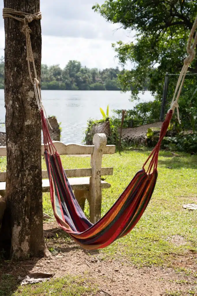 A colorful striped hammock is tied between a tree and a wooden bench near a grassy shore, overlooking a calm lake—an inviting spot to relax and unwind, perfect for those seeking peaceful things to do in Ahangama.