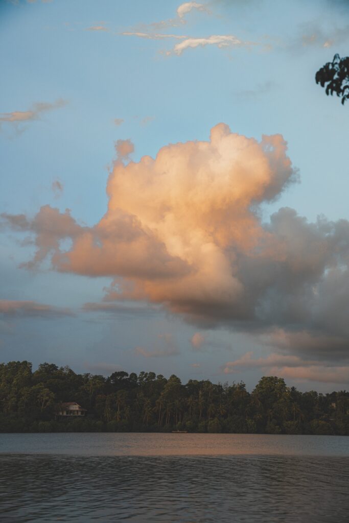 A large, fluffy cloud glows with warm sunlight in the sky above a calm lake, with trees and a small house along the shorelineโinviting you to relax and imagine peaceful things to do in Ahangama.