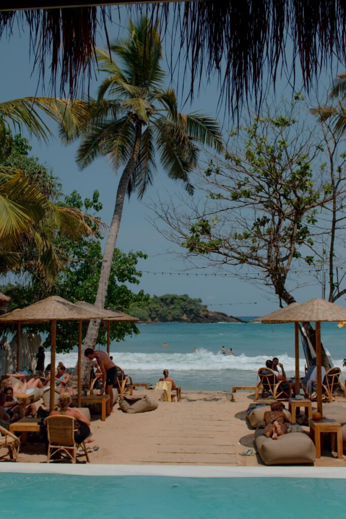 People relax on lounge chairs and bean bags under umbrellas beside a pool, overlooking a tropical beach with palm trees and blue ocean waves—one of the top things to do in Hiriketiya for a laid-back coastal escape.