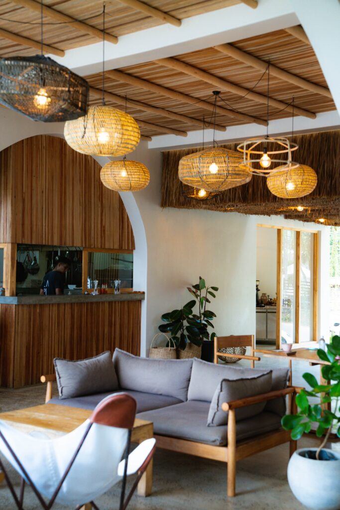 A modern, cozy café interior with wooden and rattan accents, hanging woven lights, a gray sofa, potted plants, a wooden table, and large windows letting in natural light—perfect for relaxing while exploring Things To Do In Hiriketiya.