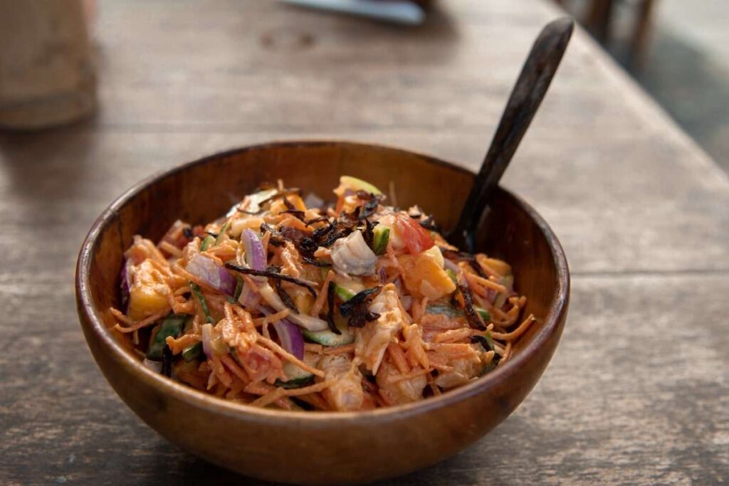 A wooden bowl filled with a colorful salad featuring shredded vegetables, red onions, seaweed strips, and creamy dressing sits on a rustic wooden table—perfect for those exploring Things To Do In Hiriketiya.