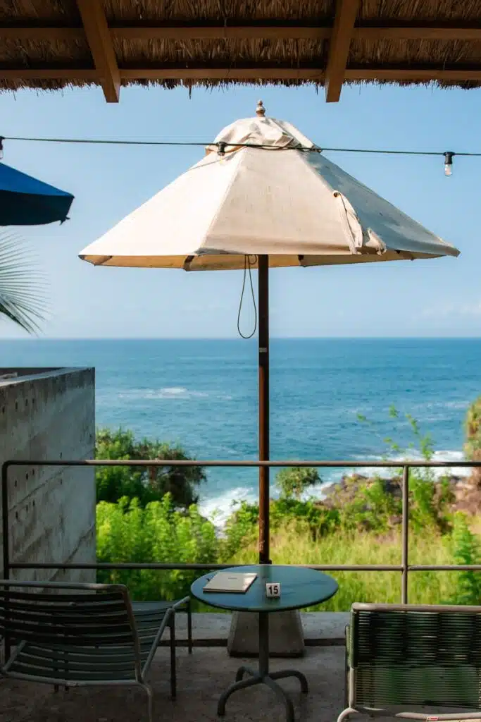 A white patio umbrella shades a small round table and chairs on a terrace overlooking the ocean, perfect for relaxing after exploring Things To Do In Hiriketiya. A booklet and table number 15 sit atop the table amidst lush greenery and blue sky.