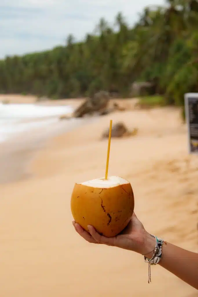 A hand holding a fresh coconut with a straw on a sandy beach, tropical trees and ocean waves in the background—one of the must-try things to do in Hiriketiya.