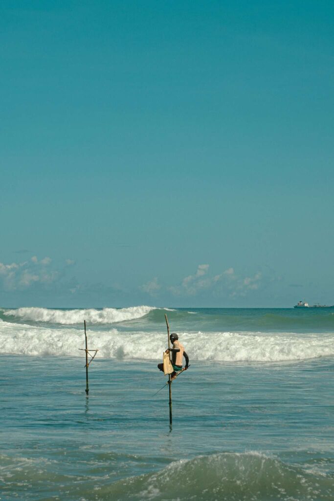 A person sits on a stilt above ocean waves, fishing in the sea under a clear blue skyโone of the unique things to do in Ahangama. Another empty stilt stands nearby as waves crash toward the shore in the background.