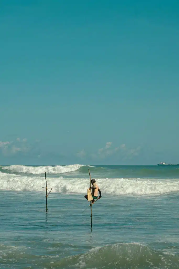 A person sits on a stilt above ocean waves, fishing in the sea under a clear blue sky—one of the unique things to do in Ahangama. Another empty stilt stands nearby as waves crash toward the shore in the background.