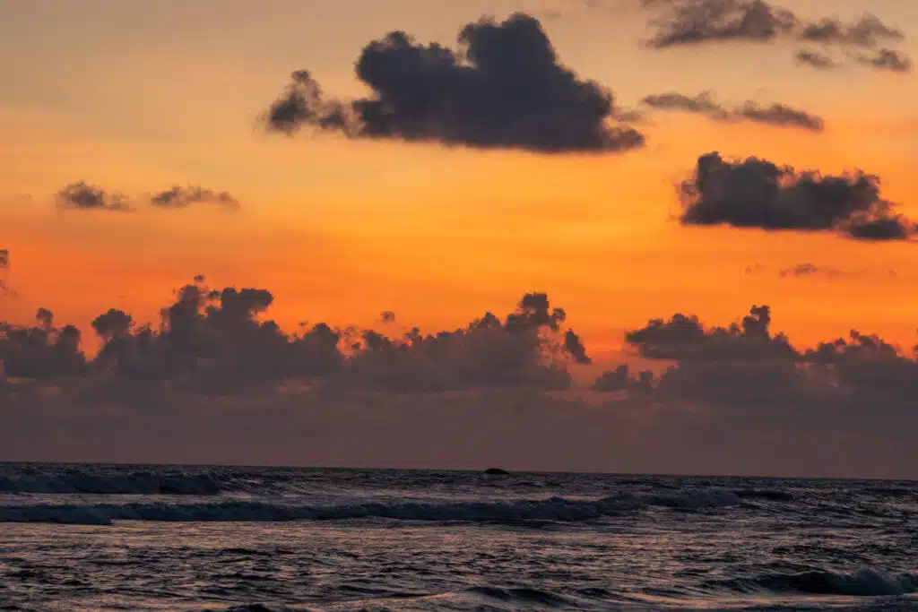 Waves gently roll onto the sea under a vibrant orange sunset sky, scattered with dark clouds. The horizon separates illuminated clouds from the darker water below—one of many breathtaking scenes among things to do in Ahangama.