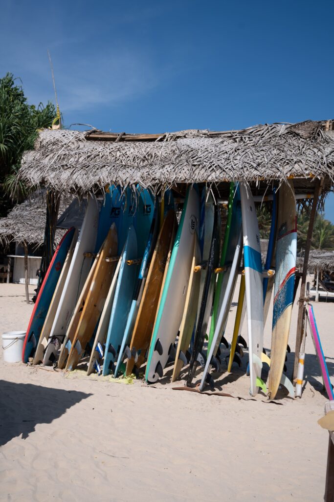 A row of colorful surfboards is propped upright under a rustic palm-thatched hut on a sandy beach, showcasing one of the best things to do in Ahangama, with clear blue skies and tropical greenery in the background.