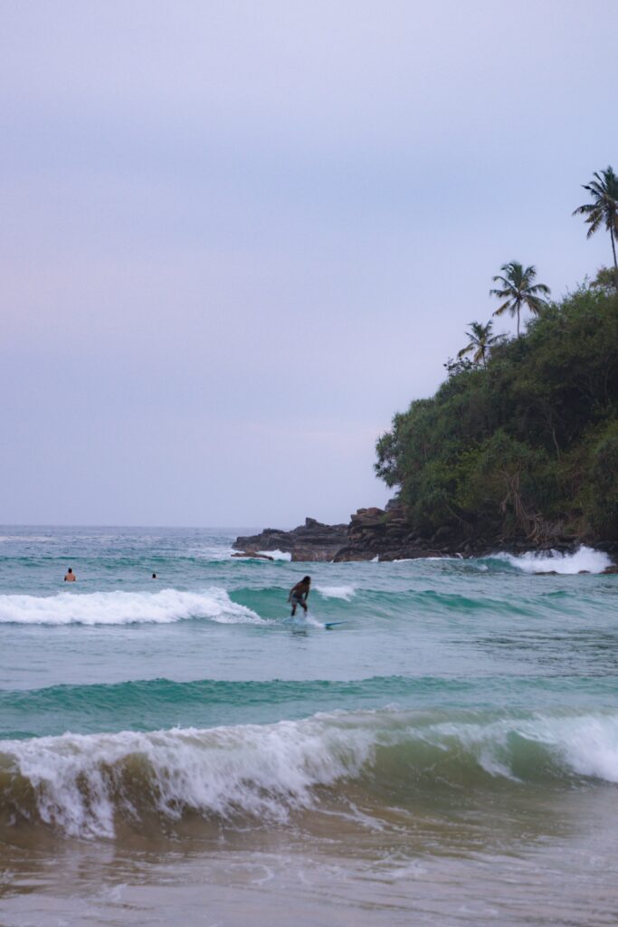 A surfer rides a gentle wave near Hiriketiya’s rocky, palm-fringed shore under a cloudy sky—a classic scene among the top things to do in Hiriketiya. Turquoise waters and other surfers complete this serene coastal moment.