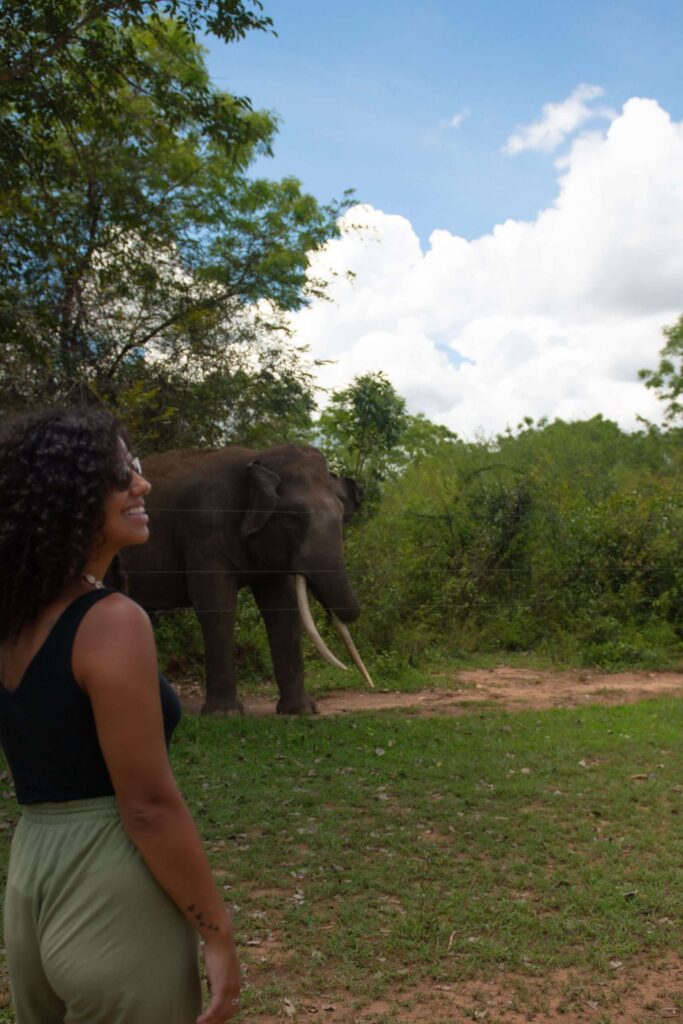A woman with curly hair stands smiling in the foreground, while an elephant with long tusks walks behind her in a lush, green landscapeโone of the unforgettable things to do in Ahangama under a partly cloudy sky.
