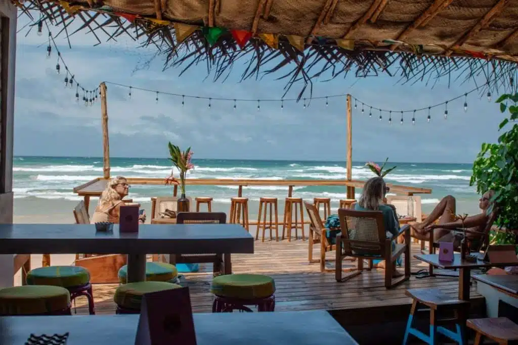 A beachfront café with wooden tables and colorful stools overlooks the ocean, embodying the laid-back charm typical of restaurants in Ahangama. String lights hang overhead as patrons sit and relax, savoring the view of the waves under a cloudy sky.