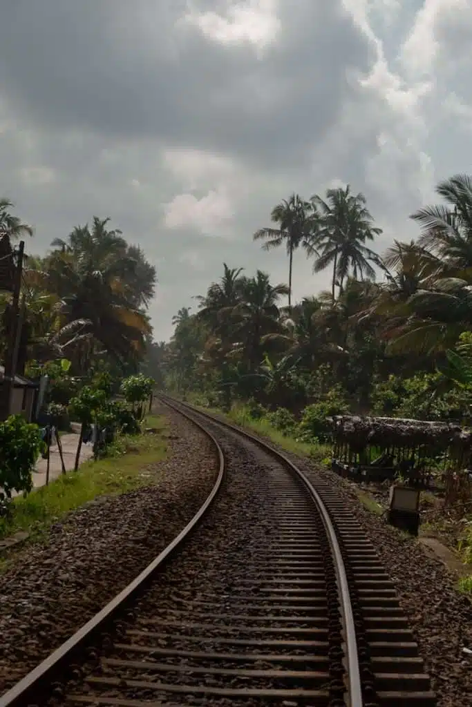 A curved railway track weaves through a tropical landscape near Ahangama, flanked by palm trees under a cloudy sky. On the left, a small road parallels the tracks, inviting exploration amid lush greenery.