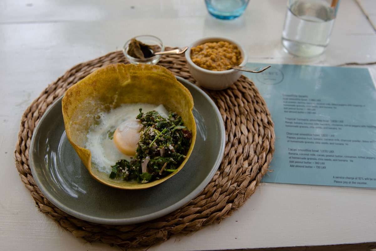 A plate showcasing a unique dish with a fried egg and greens in a crispy shell sits beside a bowl of granola and a jar of dark condiment, all elegantly presented on a woven placemat. Perfect for fueling your adventures in Hiriketiya, where endless things to do await.