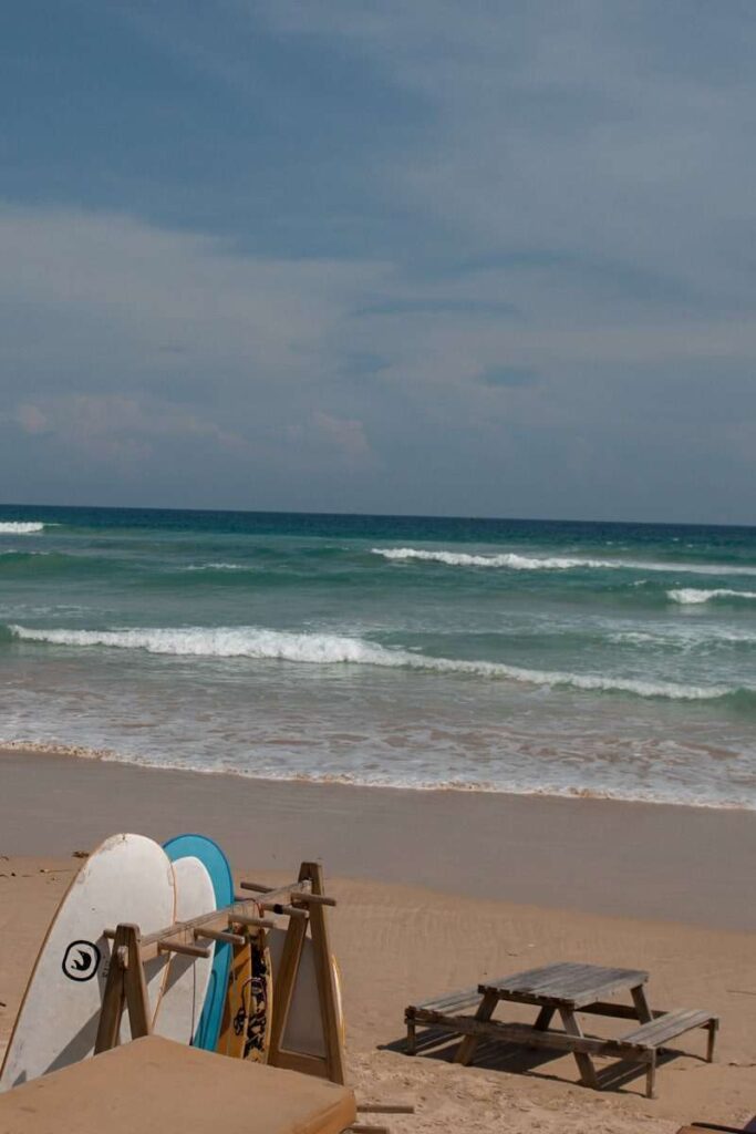 Three surfboards rest against wooden stands on a sandy beach with a picnic table nearby. Gentle waves roll onto the shore under a partly cloudy skyโperfect for those seeking things to do in Ahangama.