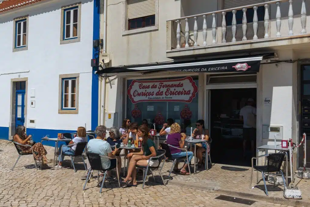 People sit at outdoor tables enjoying food and drinks in front of a bakery called "Casa da Fernanda Ouriços da Ericeira," one of the Best Restaurants In Ericeira, on a sunny day in a cobblestone street, with white buildings in the background.