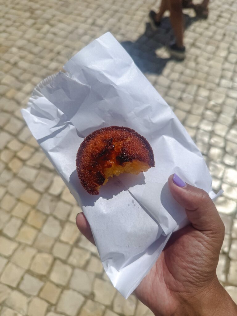 A hand with lavender nail polish holds a half-eaten pastry in a white paper bag, with a cobblestone street and pedestrians in the background—an everyday scene near some of the Best Restaurants In Ericeira.