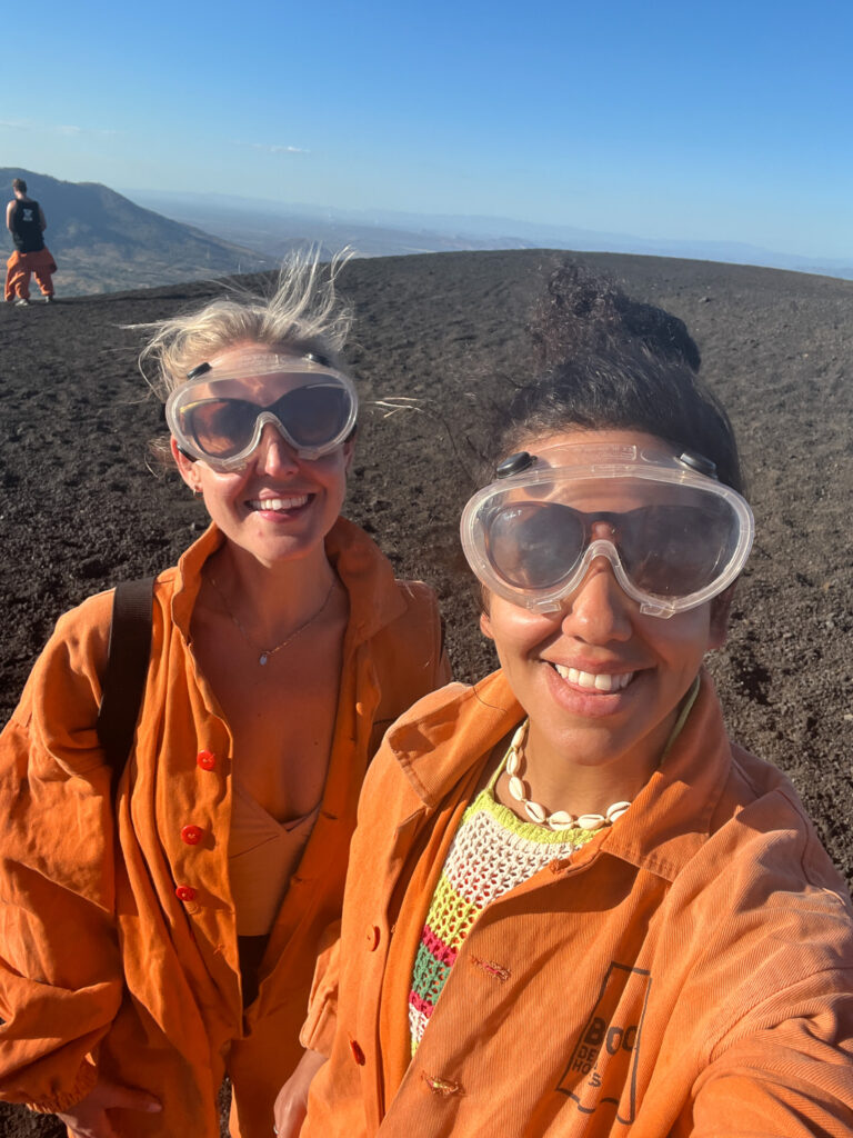 Two women in orange jumpsuits and large protective goggles smile outdoors on a dark, rocky landscape beneath a clear blue sky—just one adventure from a 10-Day Nicaragua Itinerary. A third person stands in the background near distant hills.