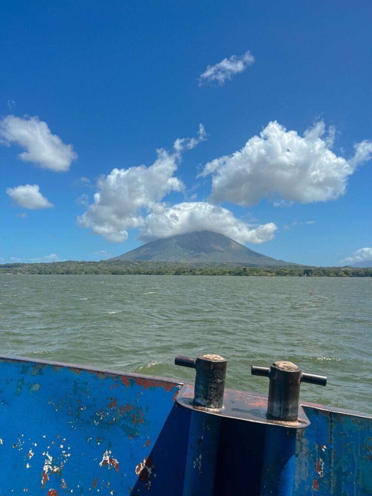 A blue, weathered boat railing frames the water and a distant volcano shrouded in clouds beneath a bright sky. Lush trees line the shore—an unforgettable sight on any 10-Day Nicaragua Itinerary.