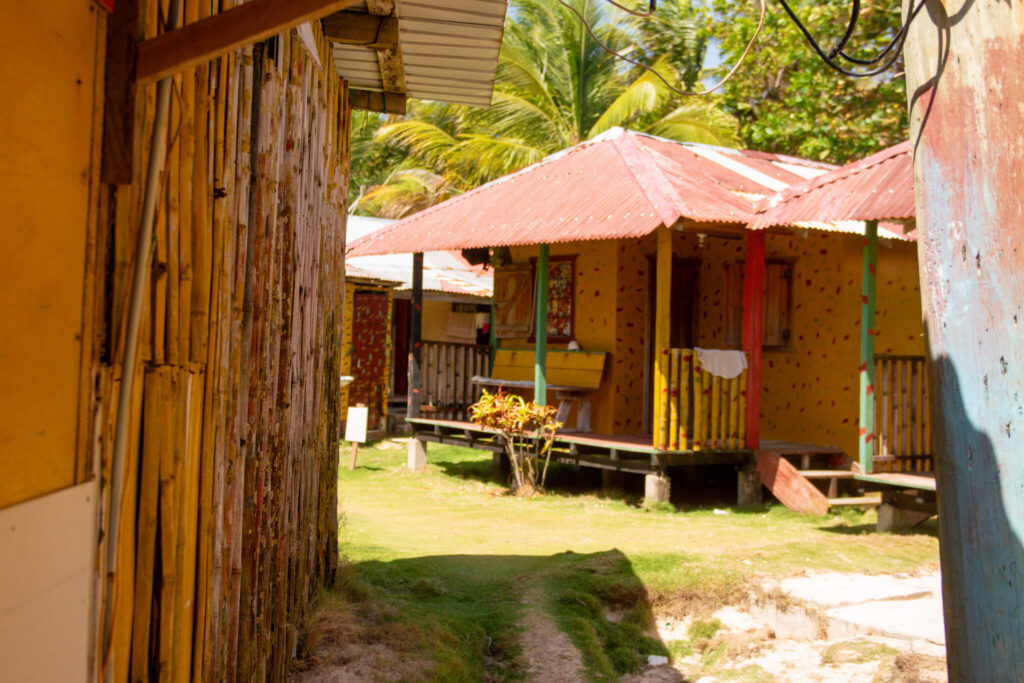 Colorful wooden houses with red tin roofs sit on a grassy yard lined with palm trees; a bamboo fence and a weathered blue wall stand in the foreground, capturing the charm found along a 10-Day Nicaragua Itinerary on this sunny day.