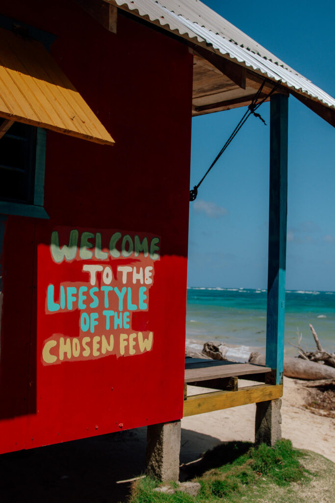 A bright red beach hut with a painted sign reading "Welcome to the lifestyle of the chosen few" stands on sandy ground near turquoise ocean water—an inviting stop on your 10-Day Nicaragua Itinerary under a clear blue sky.