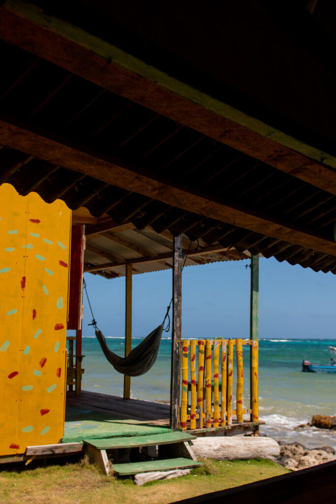 A colorful beach hut with a yellow door and bamboo railing sits on the shore—perfect for a relaxing stop on your 10-Day Nicaragua Itinerary. A hammock hangs under the roof, facing the ocean, with blue sky and waves in the background.