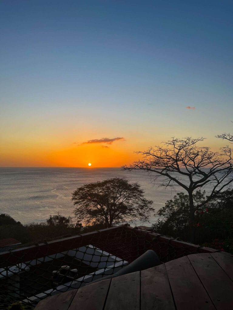 A vibrant sunset over the ocean with an orange sky, silhouetted trees in the foreground, and rooftops partially visible at the bottom—an ideal scene from a 10-Day Nicaragua Itinerary.